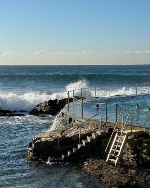 Ocean pool in Australia with people swimming on a sunny day. Waves are crashing into the pool barrier as people swim in the pool.