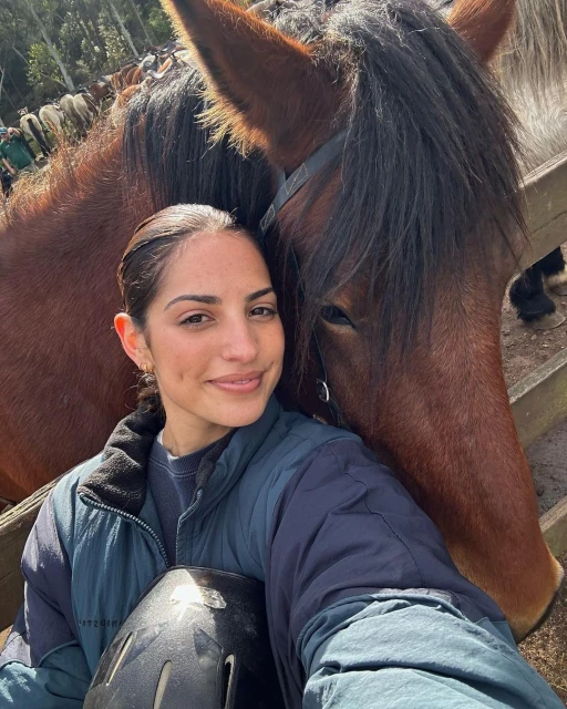 Woman poses for a selfie with a brown horse outdoors during the day. Trees are visible in the background.