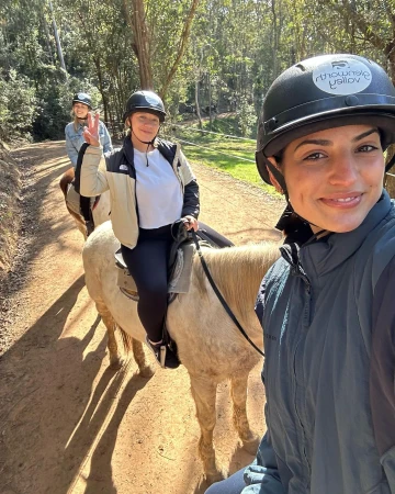 Three women ride horses on a dirt path lined with trees; the woman in front holds the camera at eye level for a selfie.