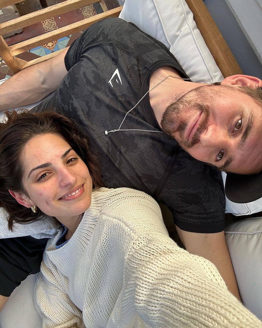 High-angle selfie of a smiling woman in a cream sweater and a man in a black patterned t-shirt relaxing on cushions.