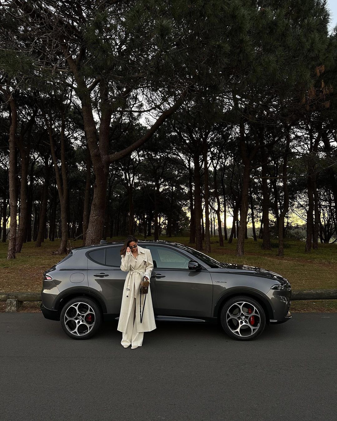 Woman posing next to a gray SUV in an outdoor setting with trees, wearing a light-colored trench coat and pants, holding a purse.