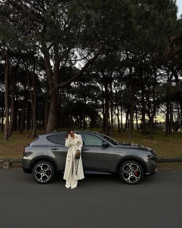 Woman in white coat standing beside a dark grey SUV parked on an asphalt road framed by tall trees.