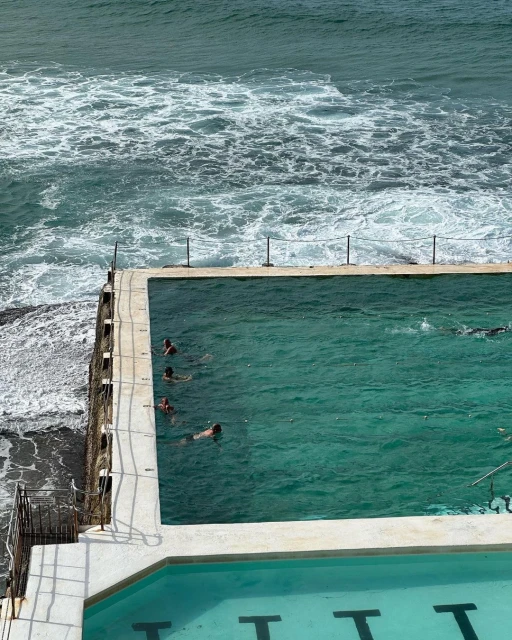 People swim in an ocean-fed saltwater pool with a rocky coastline. Ocean waves crash against the pool’s edge on a sunny day.