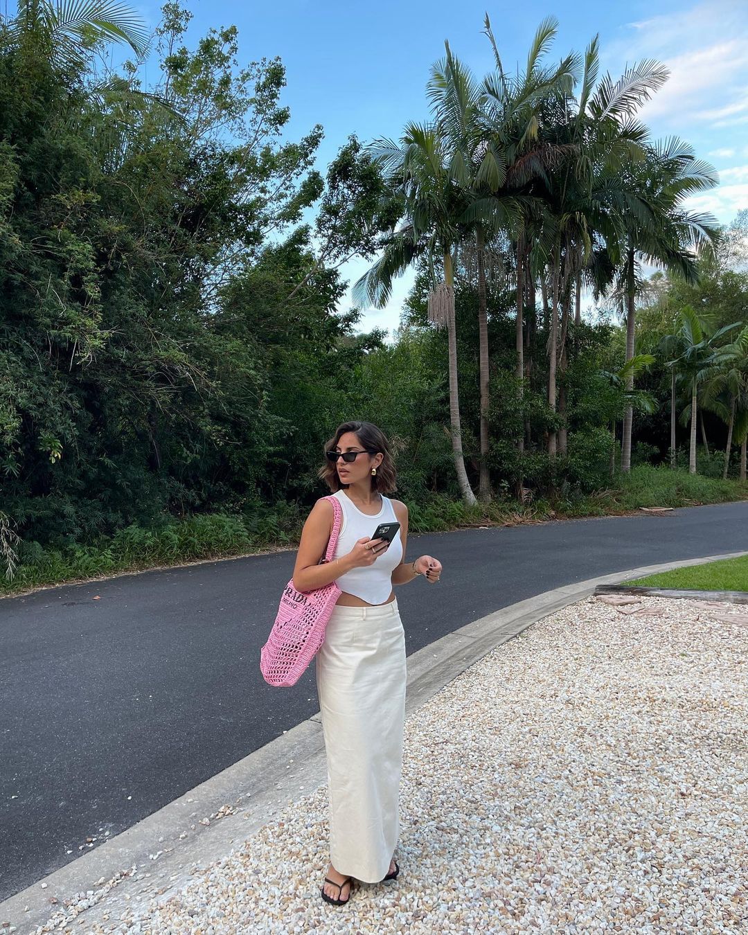 Woman wearing white top and skirt holds phone on a sunny day, pink crocheted bag on shoulder, palm trees and plants in background.
