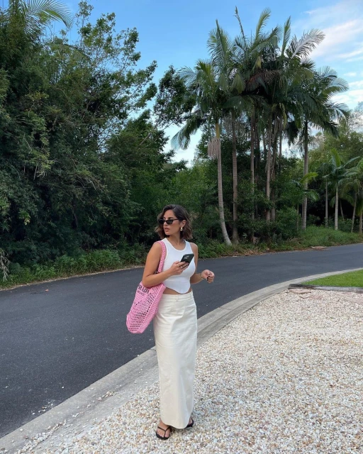 Woman wearing white top and skirt holds phone on a sunny day, pink crocheted bag on shoulder, palm trees and plants in background.