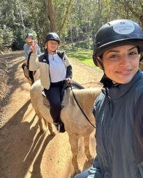 Three smiling women wearing riding helmets on horseback along a sunny dirt forest trail