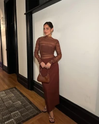 Woman in brown mesh top and brown skirt holding a small handbag standing in a hallway
