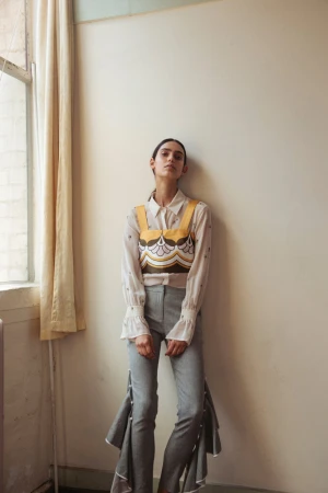 Model leaning against a neutral wall wearing patterned top over white blouse and grey trousers styled by Jade Leung