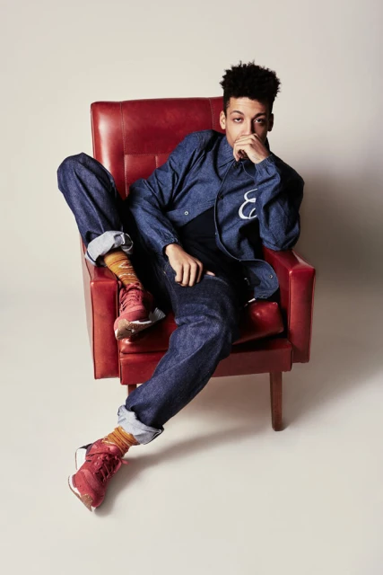 An adult male in a denim outfit poses in a red armchair with red shoes, against a solid background in a studio photography shot.