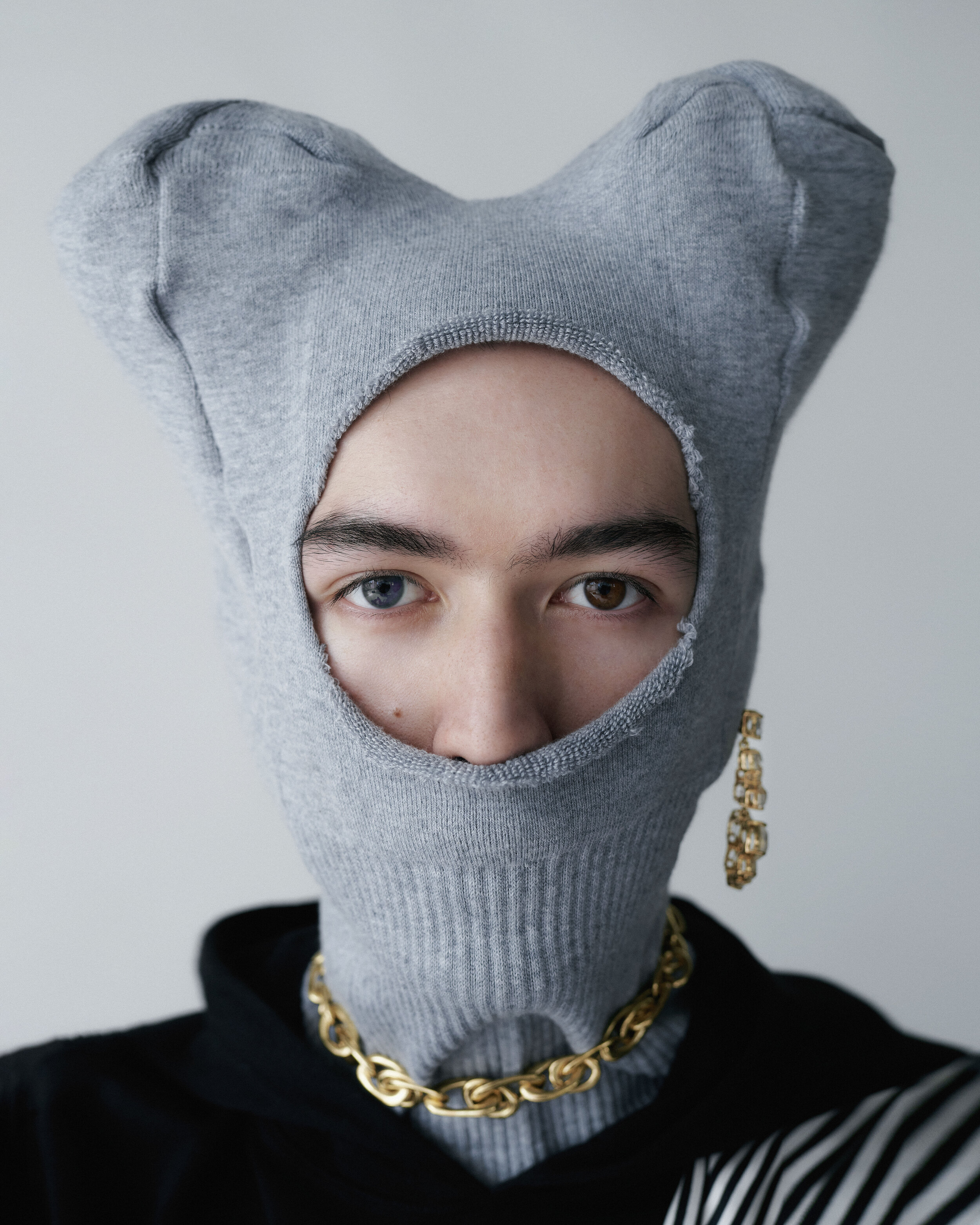 Close-up portrait of a person wearing a gray balaclava, gold chain necklace, and dangling gold earrings against a neutral backdrop.