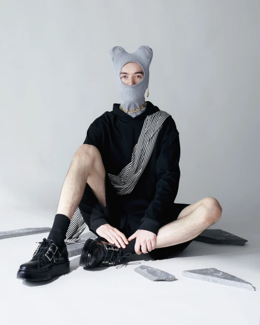 Person wearing a grey balaclava, gold chain, black hoodie, and patent leather shoes sitting on a white surface with grey stones.