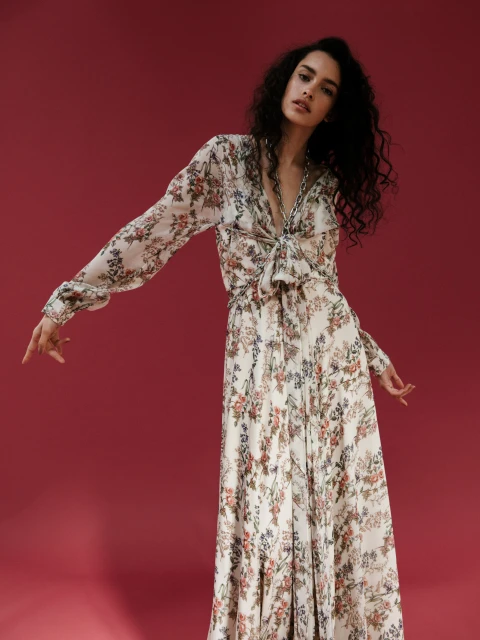A young adult woman with curly hair in a long-sleeve floral dress poses against a plain red backdrop, striking a pose with outstretched arms.