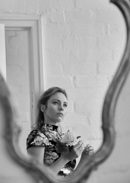 Black-and-white portrait of a blond woman holding a flower, framed by a mirror against a white brick wall with a window.