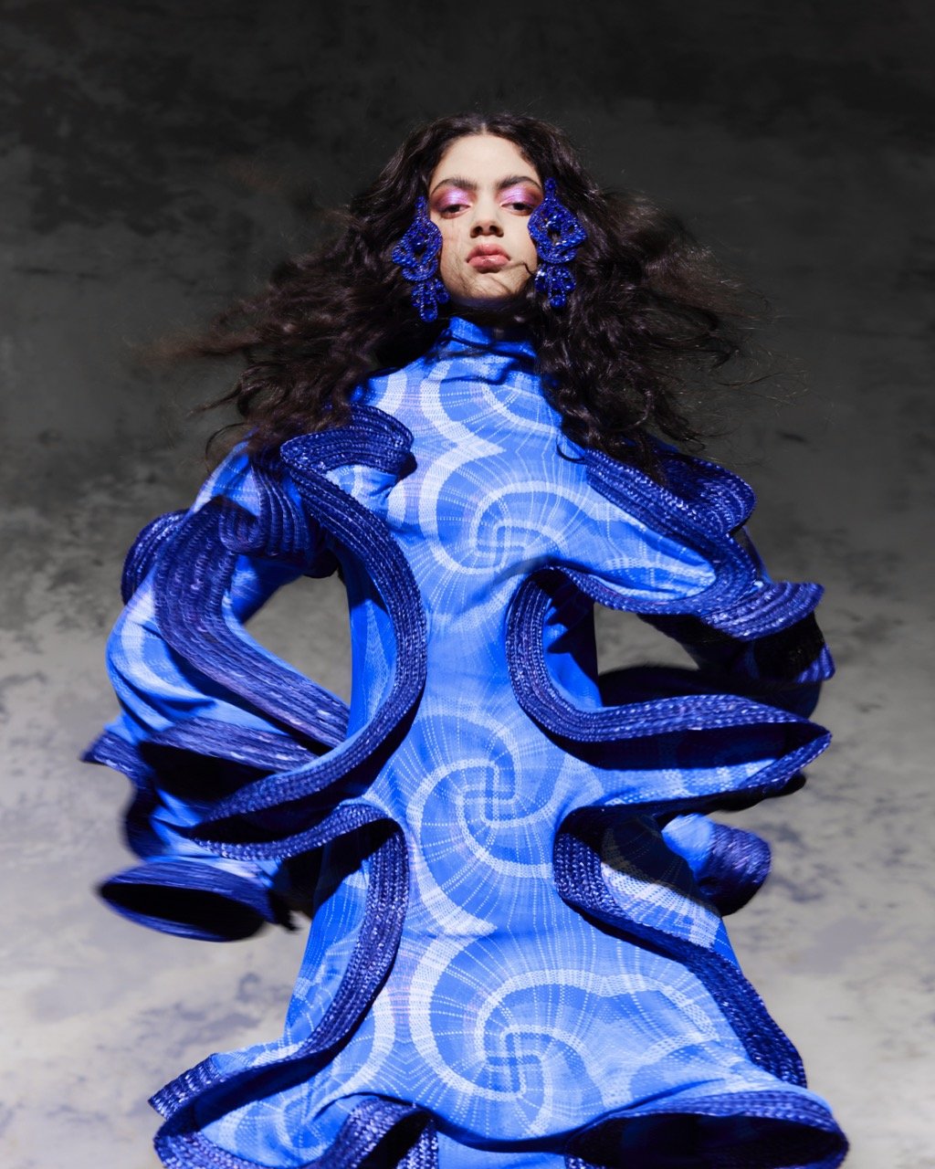 Brunette woman in a blue patterned dress with ruffled trim and blue beaded earrings, posing against a grey textured background in a studio shot.