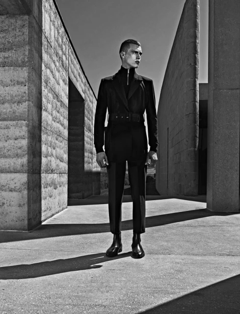 Monochrome portrait of a man in a dark suit posing outdoors against a concrete architectural background in bright, neutral lighting.