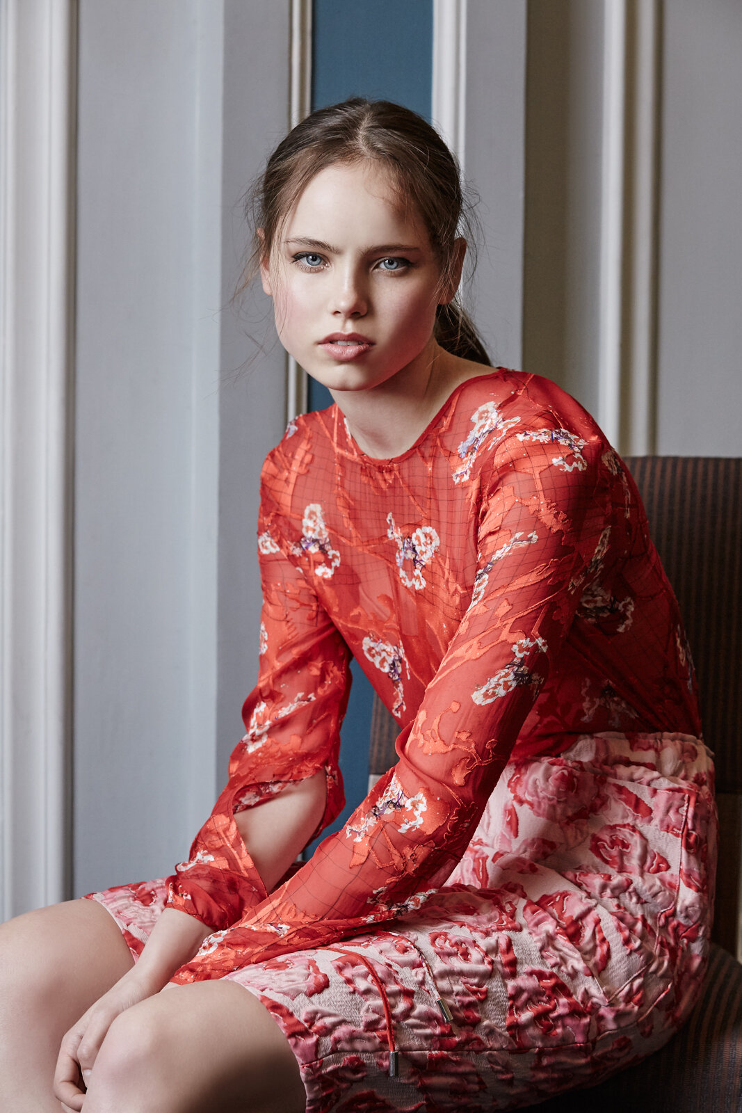 Portrait of a young adult woman with floral top and skirt sitting indoors in front of a wall with vertical stripes and moulding.