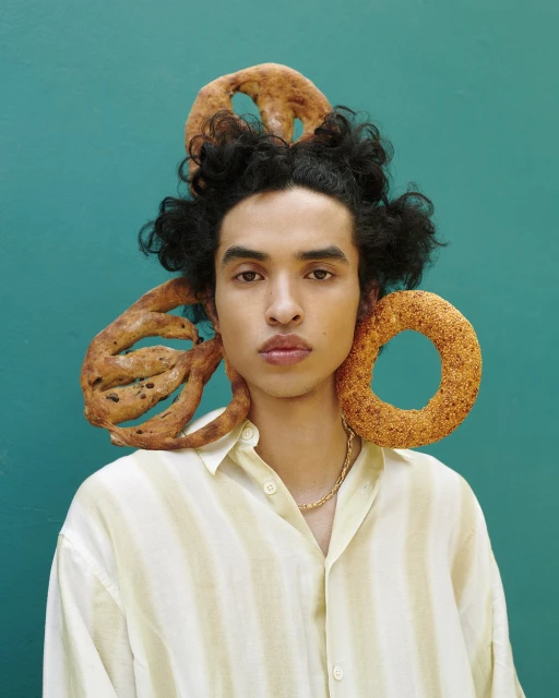 Portrait of a young adult with curly hair wearing a striped shirt, adorned with bagels and standing against a teal background.
