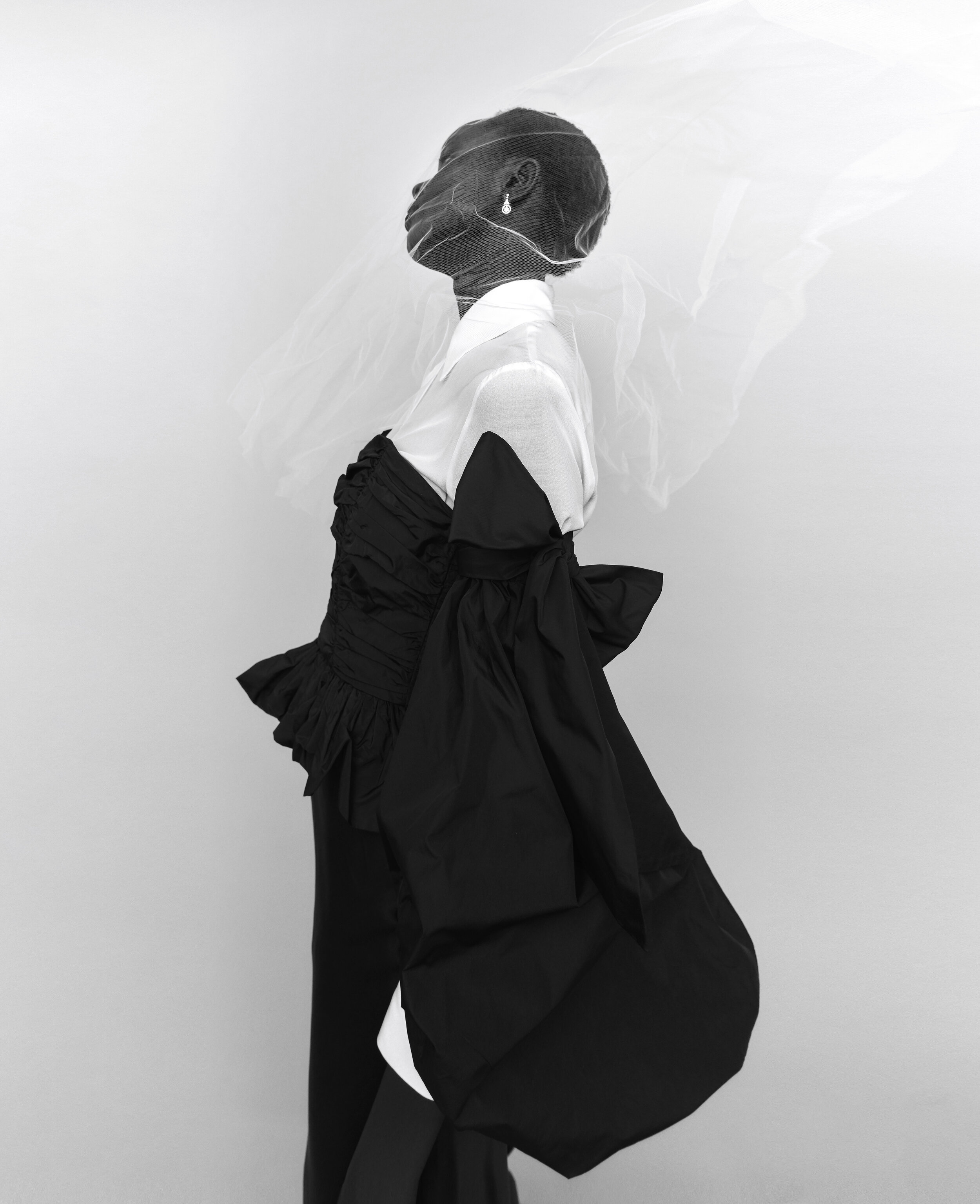 Elegant portrait of a dark-skinned woman wearing a black ruffled dress with a white shirt, sheer veil, and pearl earring against light grey backdrop.
