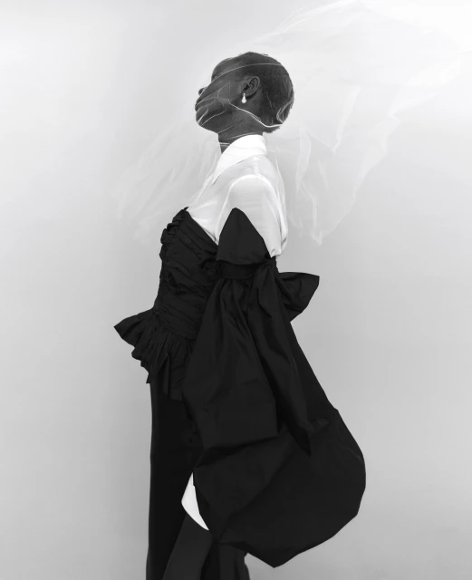 Elegant portrait of a dark-skinned woman wearing a black ruffled dress with a white shirt, sheer veil, and pearl earring against light grey backdrop.