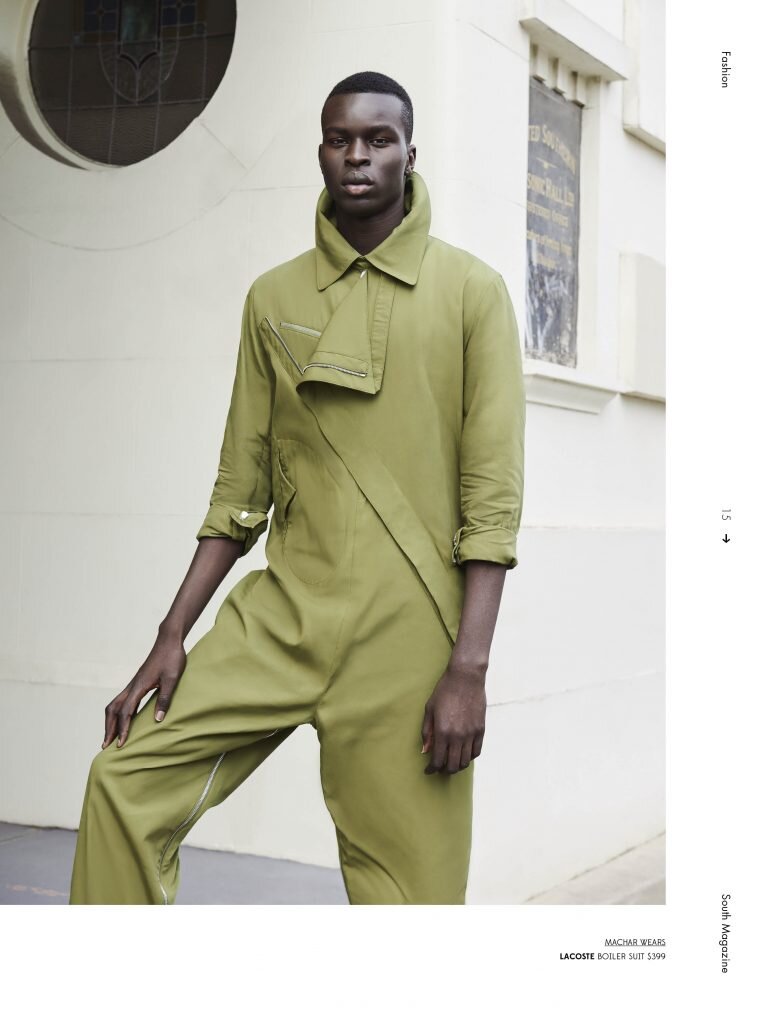 Fashion editorial portrait of a Black male model in an olive green jumpsuit, posing against a light-colored building with architectural details.