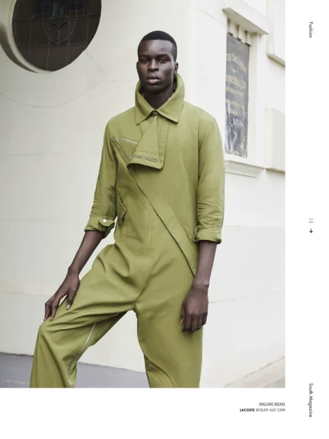 Fashion editorial portrait of a Black male model in an olive green jumpsuit, posing against a light-colored building with architectural details.