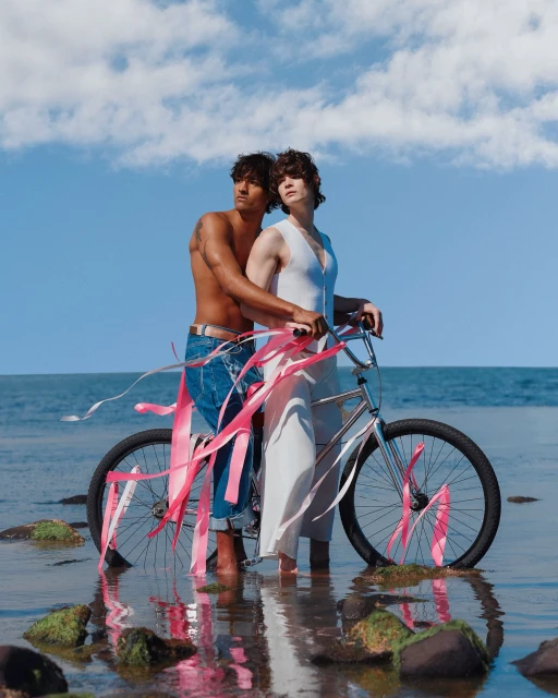 Two shirtless adults with curly dark hair pose in the ocean with a silver bicycle decorated with pink ribbons under a partly cloudy blue sky.