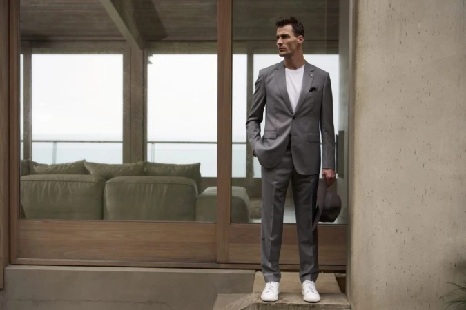 Man in a grey suit and white t-shirt standing outside modern architecture holding a hat, styled by Jade Leung
