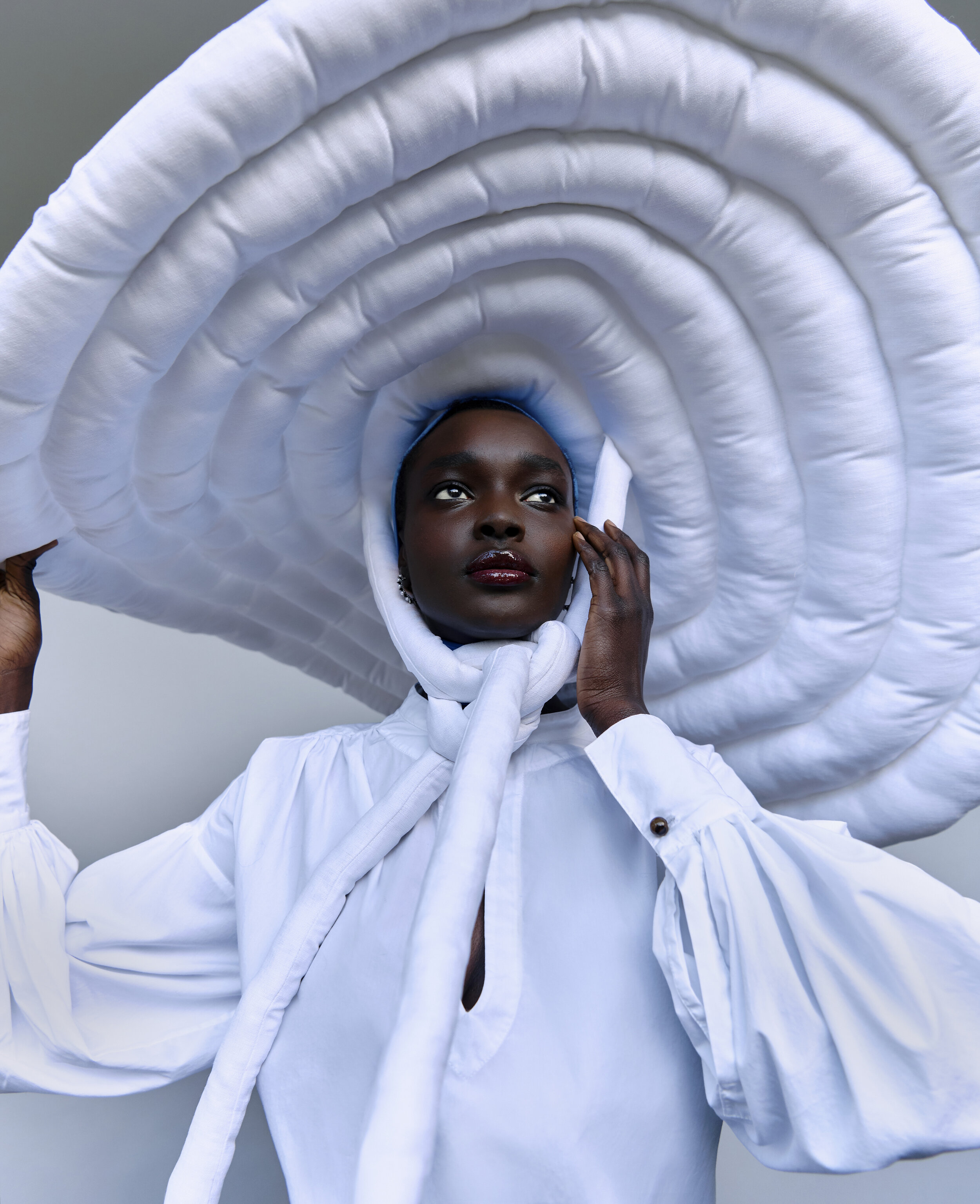 An adult Black woman with dark skin poses in a white top and a dramatic, sculptural white hat. Bright, diffused light illuminates the studio.