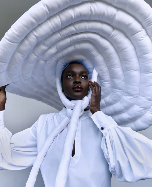 An adult Black woman with dark skin poses in a white top and a dramatic, sculptural white hat. Bright, diffused light illuminates the studio.