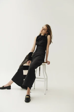 Model in black sleeveless top and trousers sitting on a white stool against a plain background