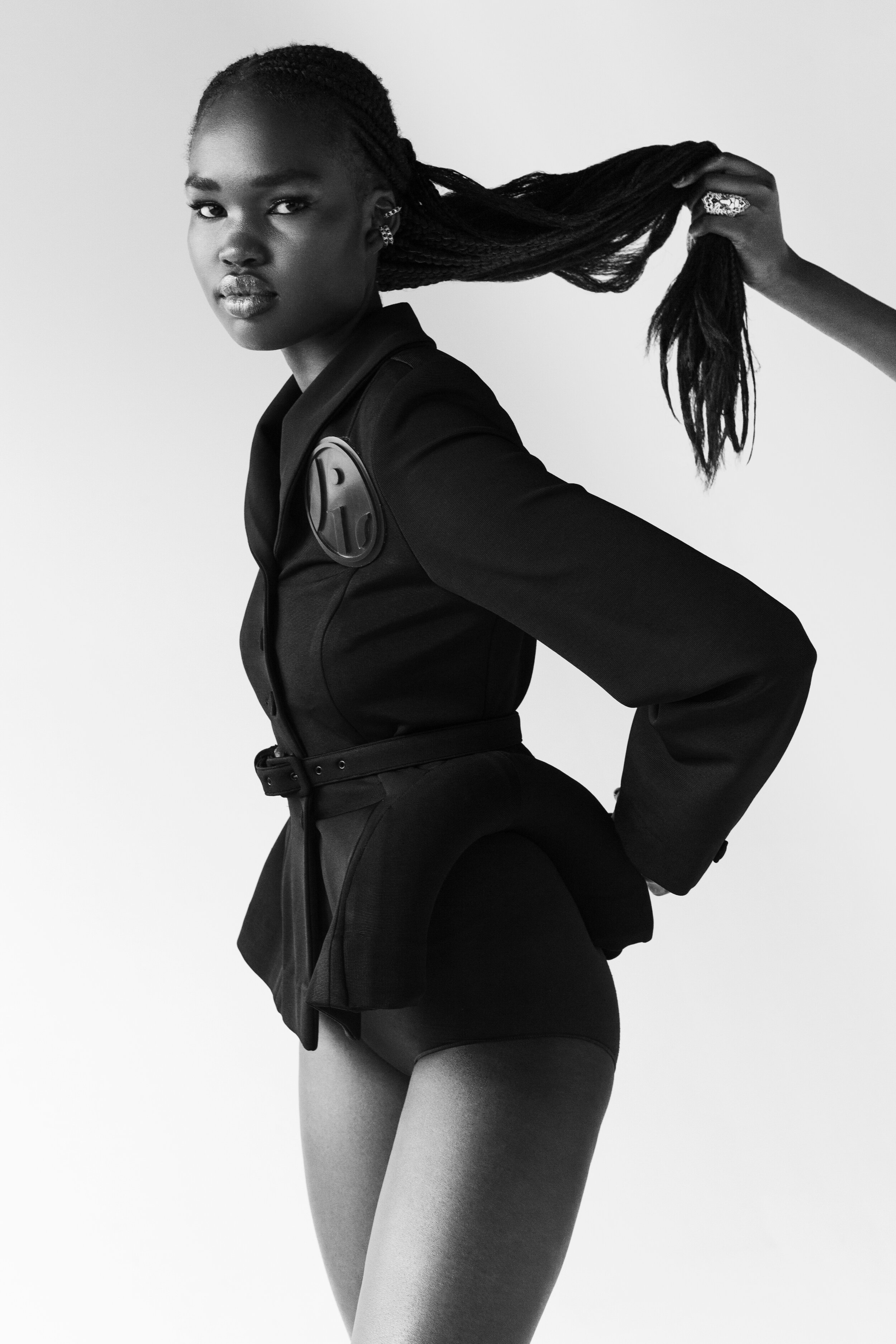 Black and white portrait of a young Black woman in a black suit, holding braided hair, against a plain backdrop in a photo studio.