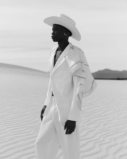 Elegant black woman in all-white outfit poses in desert landscape under bright daylight, fashion shoot, neutral monochromatic tone.
