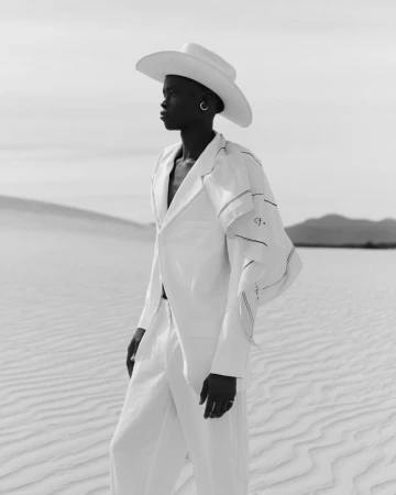Elegant black woman in all-white outfit poses in desert landscape under bright daylight, fashion shoot, neutral monochromatic tone.