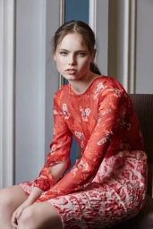 A young female model seated indoors wearing a red patterned long-sleeve top and patterned skirt styled by Jade Leung