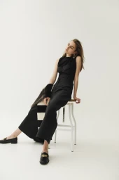Model in black sleeveless top and trousers sitting on a white stool against a plain background