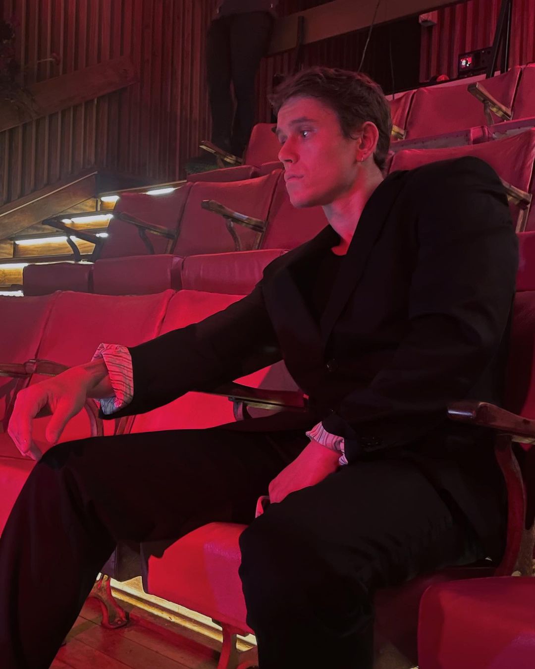 A handsome adult male in a dark suit sits in a red theatre seat. He is illuminated by red lighting in an indoor environment.