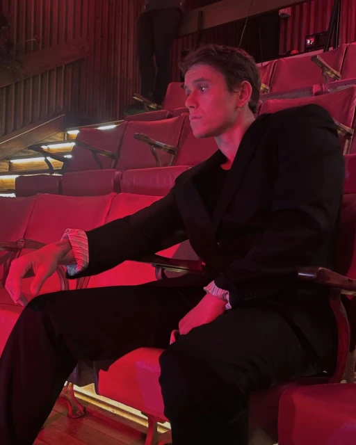 A handsome adult male in a dark suit sits in a red theatre seat. He is illuminated by red lighting in an indoor environment.