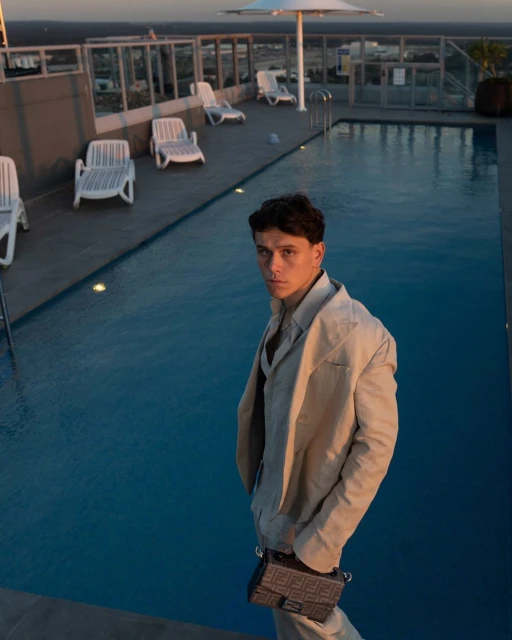 Young man in suit stands by a rooftop pool at sunset, city visible in the background, holding a dark handbag, stylish and confident.