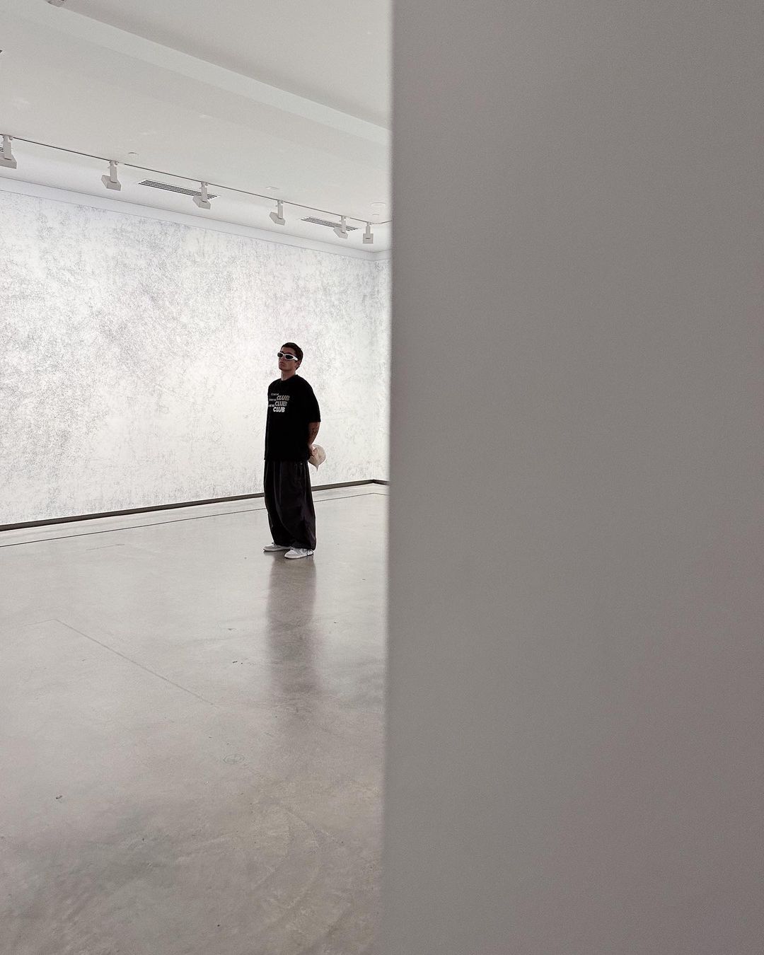 Man in sunglasses stands in a minimalist art gallery, wearing a black shirt and dark pants, contemplating the artwork on a white wall.