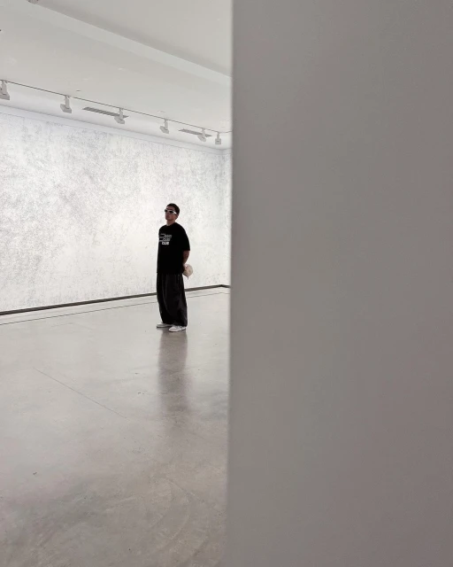 Man in sunglasses stands in a minimalist art gallery, wearing a black shirt and dark pants, contemplating the artwork on a white wall.