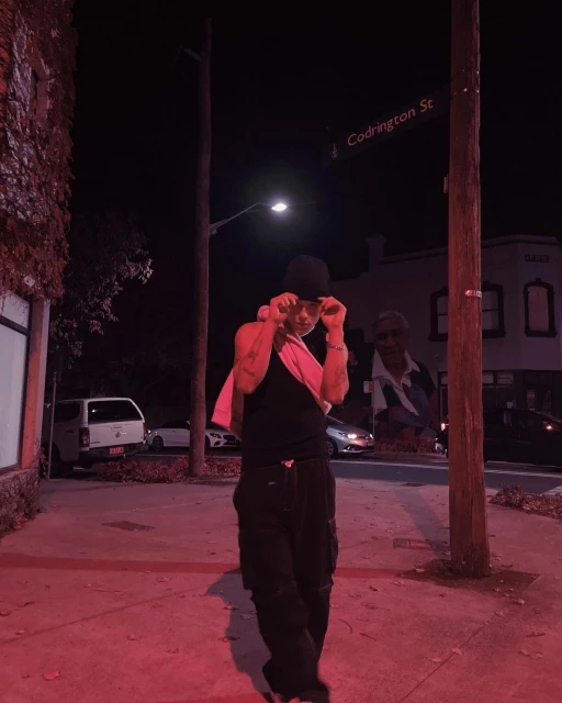 Man in a black tank top and pants poses on a city street corner at night near a street sign, with parked cars and a building visible.