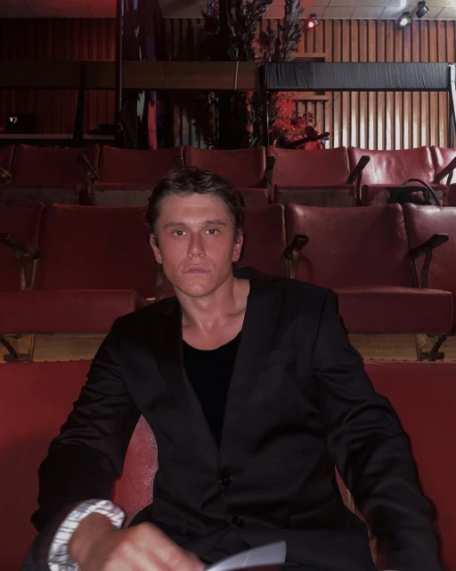 A young adult male in a black suit sits in a red theatre seat, creating a portrait-style image with a vintage aesthetic and a dimly lit atmosphere.