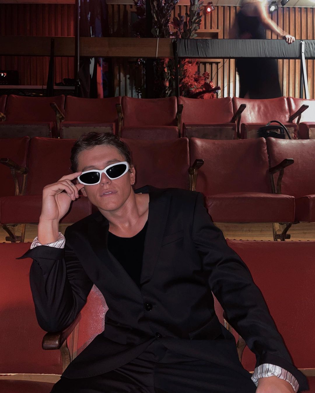 Seated man in a black suit and white sunglasses poses in a red-seated theater setting. Stylish indoor portrait. 