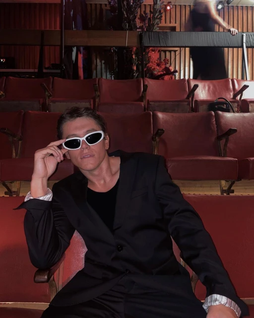 Seated man in a black suit and white sunglasses poses in a red-seated theater setting. Stylish indoor portrait. 