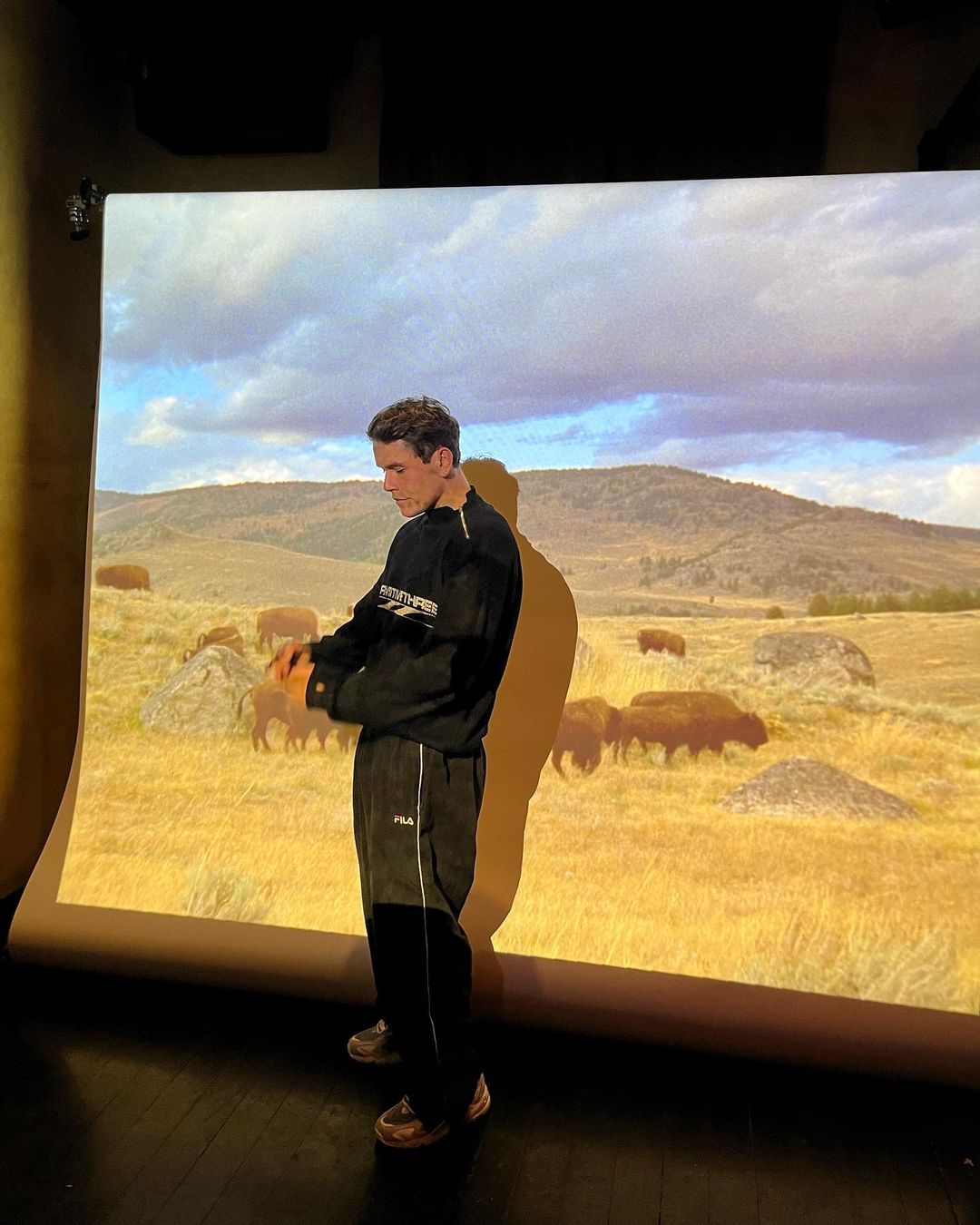 Man in a black tracksuit standing in front of a projection screen showing an image of bison in a grassy field with mountains.