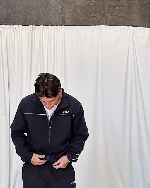 Man wearing a Fila tracksuit in front of a white curtain, casual and relaxed pose, indoor environment, neutral colors, portrait orientation.