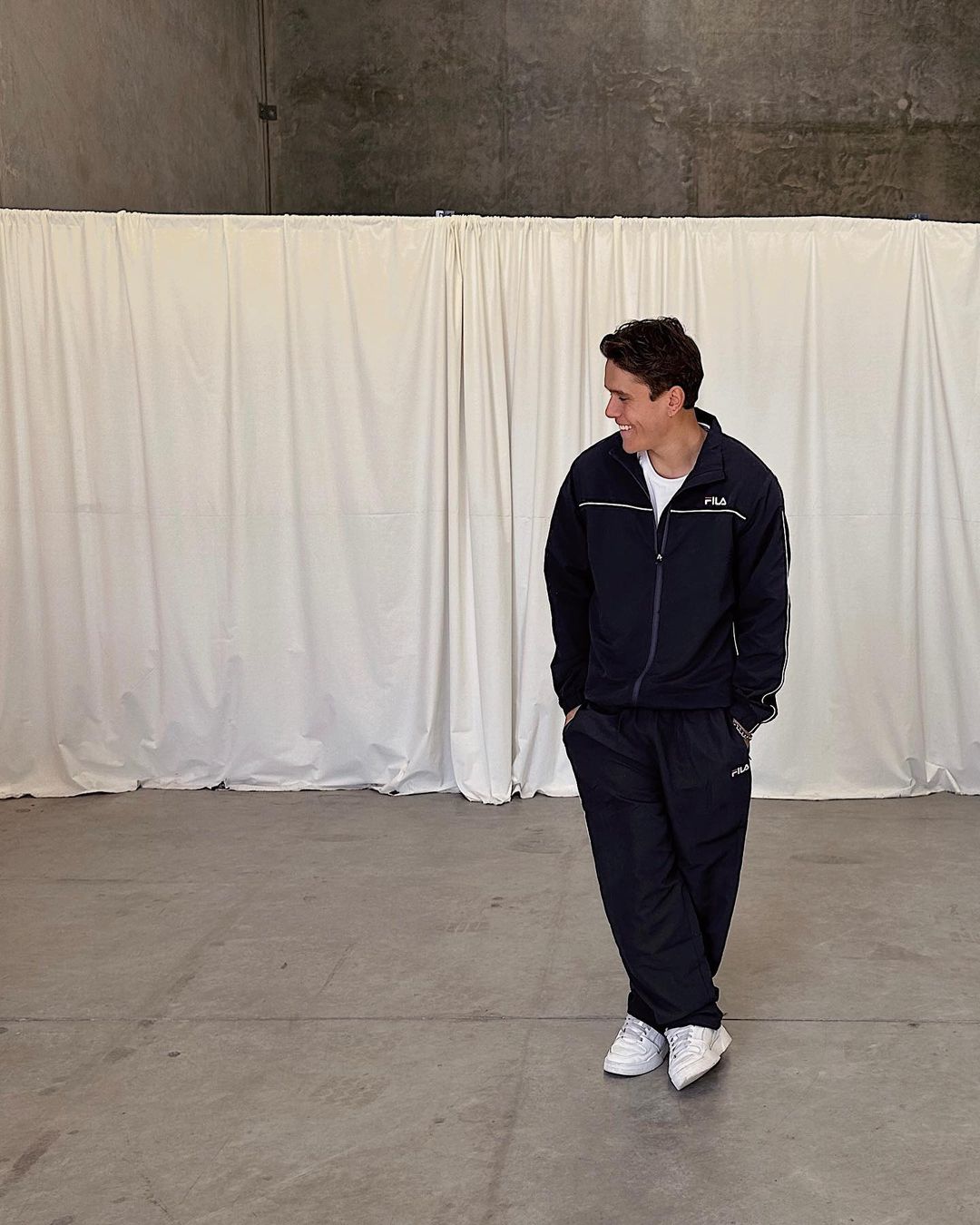 An adult male in a navy blue tracksuit stands in a minimalist indoor setting with a white curtain backdrop, looking away and smiling.