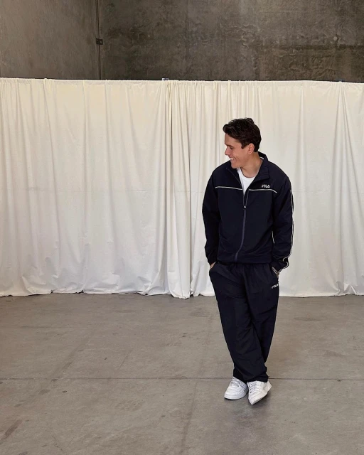 An adult male in a navy blue tracksuit stands in a minimalist indoor setting with a white curtain backdrop, looking away and smiling.