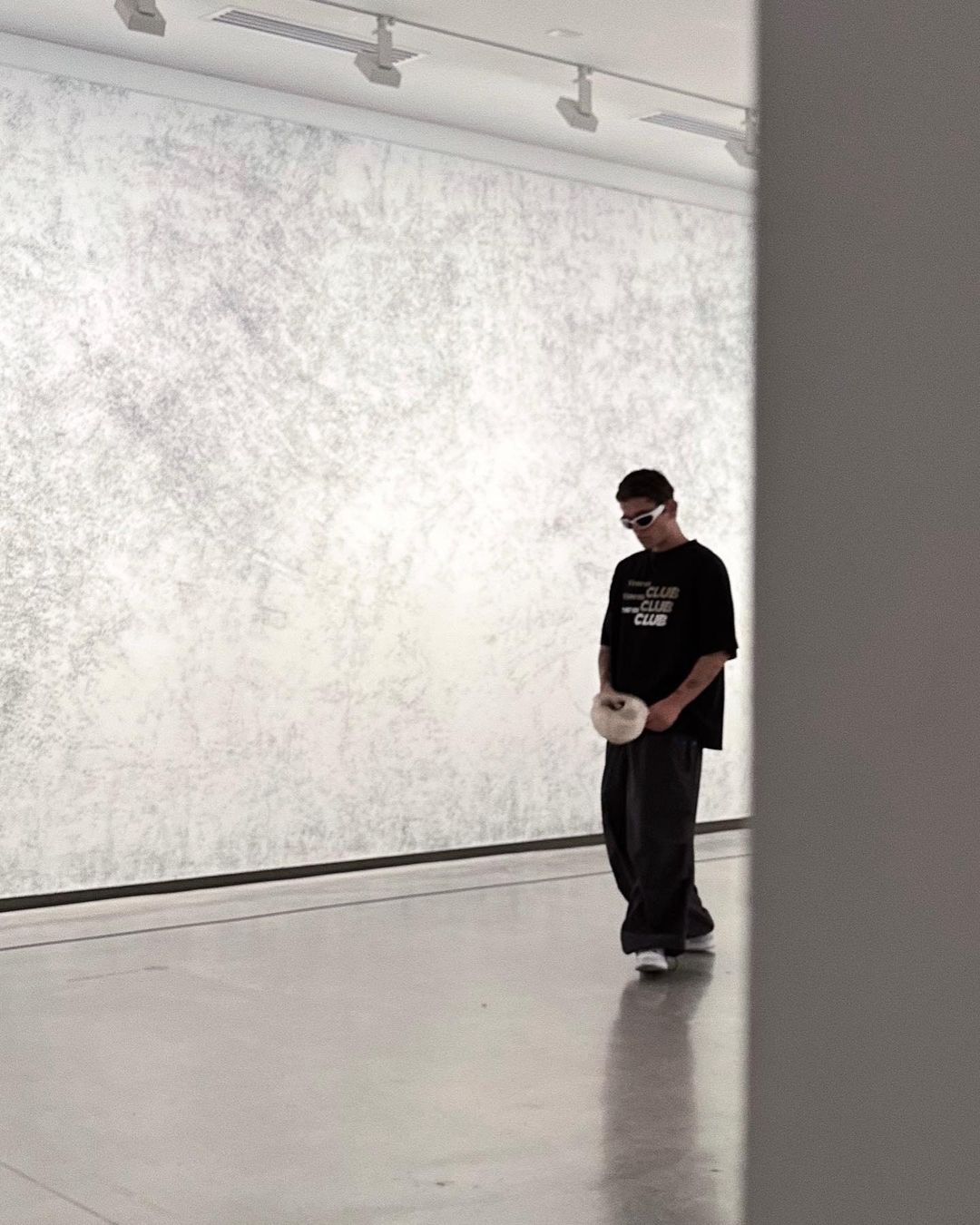 Man in casual clothing walking in a minimalist art gallery, gazing down. Neutral colors, with a textured wall and modern lighting.
