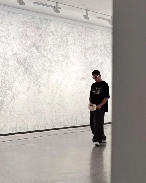 Man in casual clothing walking in a minimalist art gallery, gazing down. Neutral colors, with a textured wall and modern lighting.